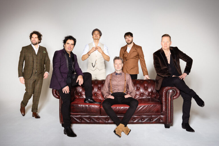 6 mannen zitten, hangen en staan om rode leren bank heen met witte achtergrond.