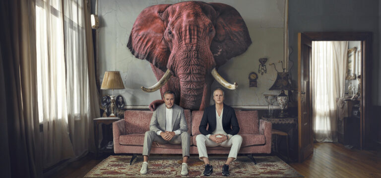 Het duo zit op een bank met draarachter een rode olifant als trofee aan de muur