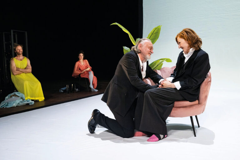 Actrice zit op roze fateuille en acteur zit geknield bij haar terwijl ze handen vasthouden. Aan de zijkant zitten andere acteurs mee te kijken.