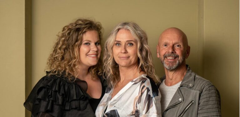 Drie mensen poseren samen voor een foto: een vrouw met halflang blond krullend haar in een zwart jurkje, een vrouw met lang grijsblond haar in een kleurrijke blouse, en een man met een baard en kort haar in een grijs jack, allemaal glimlachend naar de camera.