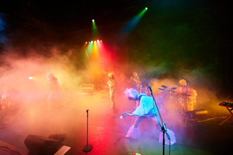 Een kleurrijk verlichte podiumscène met een groep muzikanten gekleed in theatrale kostuums en omgeven door rook, terwijl zij optreden onder felgekleurde spotlights in een energieke sfeer.