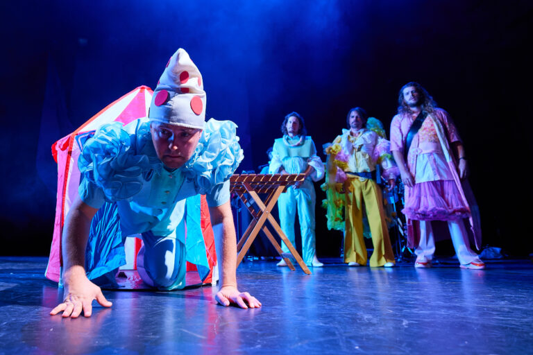Een performer in een blauw kostuum met stippenmuts kruipt op handen en knieën naar de camera op een fel verlicht podium, terwijl drie andere kleurrijk geklede artiesten op de achtergrond staan, allemaal in een theatrale setting met blauw podiumlicht.