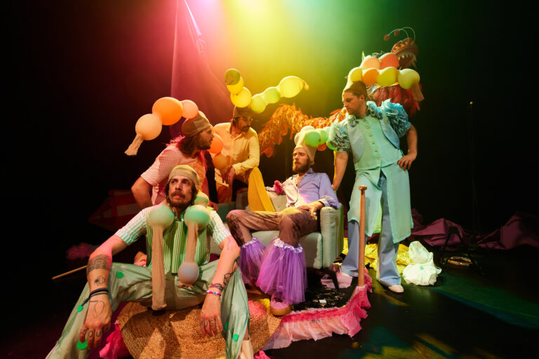 Een groep performers in extravagante, kleurrijke kostuums met ballonnen en theatrale accessoires poseren op een verlicht podium, waarbij één persoon op een stoel zit omringd door de anderen in een surrealistische scène.