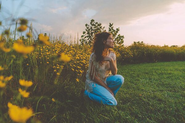 Een vrouw met een blouse en blauwe jeans zit op een groen veld, omringd door gele bloemen tijdens zonsondergang.