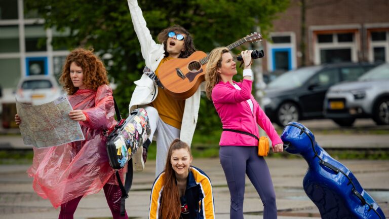 Groep kleurrijk geklede mensen die poseren; een persoon speelt gitaar terwijl anderen met accessoires in de hand staan.