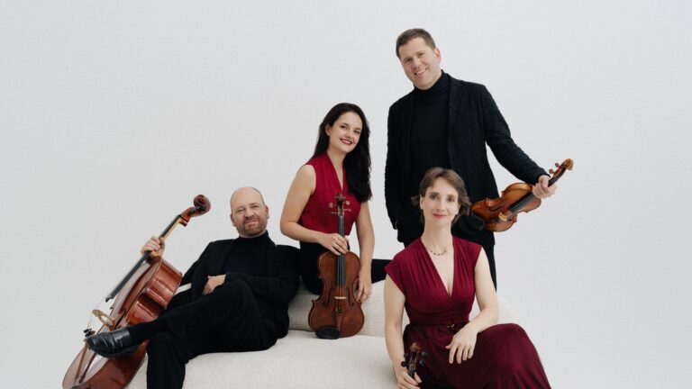 De vier musici van het strijkkwartet op witte bank tegen witte achtergrond. In het midden de twee vrouwelijke leden in het rood, geflamkeerd door de twee mannen in het zwart (de achterste man staat achter de bank)