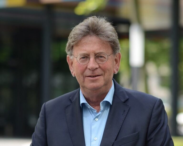 Portret van een man met grijs haar die een donkerblauw blazer en een lichtblauw overhemd draagt, staand buiten.