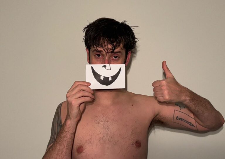 Een man met nat haar houdt een kaart met een lachend gezicht voor zijn mond en steekt zijn duim op.