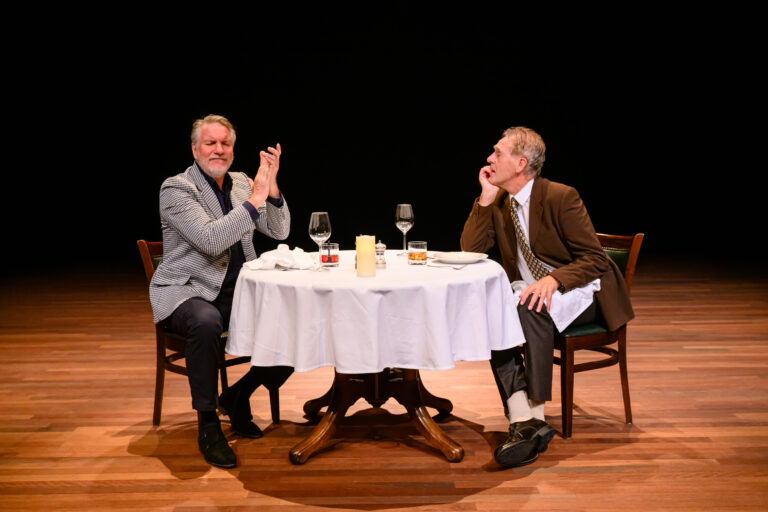 Twee mannen zitten aan een gedekte tafel met glazen wijn en kaarslicht, terwijl ze elkaar aankijken en in gesprek lijken.