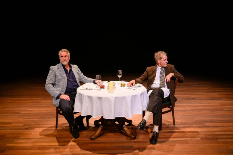 Twee mannen zitten aan een gedekte tafel met glazen wijn en kaarslicht, terwijl ze allebei expres niet naar elkaar kijken