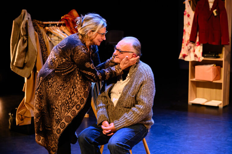 Een oudere man zit op een stoel terwijl een vrouw liefdevol zijn gezicht vasthoudt in een intieme scène, met op de achtergrond kledingrekken en een kast in een theatervoorstelling.