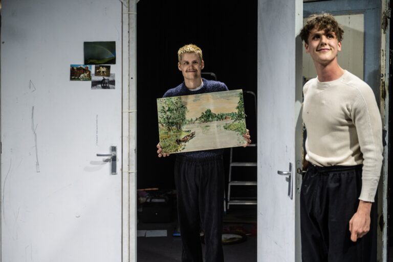 Twee mannen staan in een deuropening, waarvan één een landschapsschilderij vasthoudt terwijl de ander trots naast hem staat.