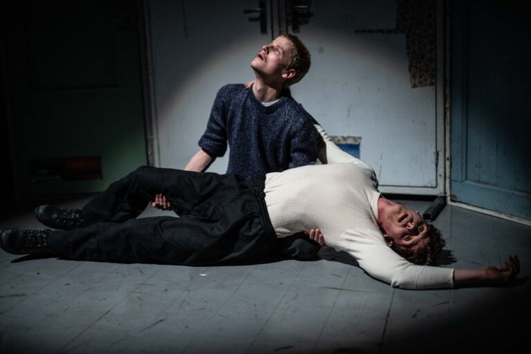 Twee mannen zitten op de grond; de ene houdt de andere, die er versuft uitziet, vast terwijl hij naar het plafond kijkt.