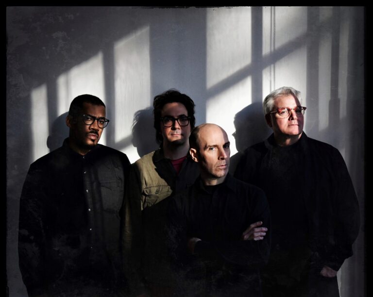 Vier mannen staan naast elkaar in een kamer met schaduwen van ramen op de muur achter hen.