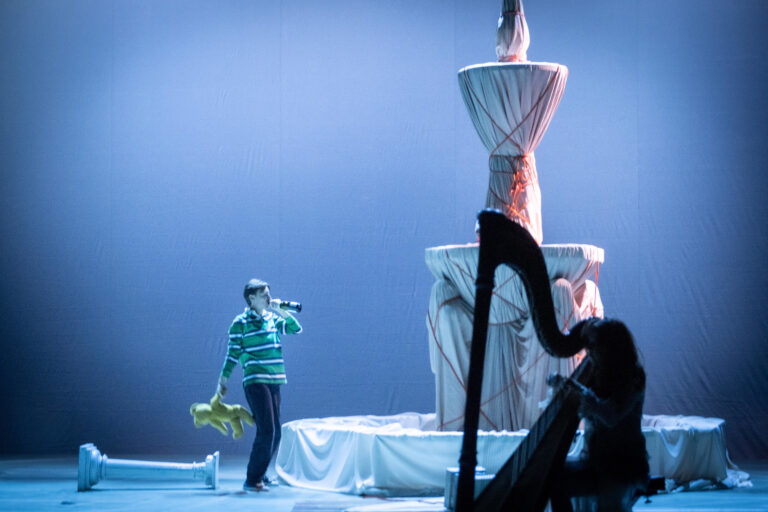 Acteur drinkt uit een drank fles, terwijl die een knuffel een diens hand heeft, en staat naast een bedoekte fontein