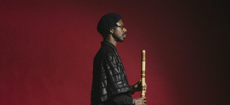 Een man in een zwarte gewaad en een zwarte baret staat in profiel tegen een rode achtergrond, terwijl hij een traditionele houten fluit vasthoudt.