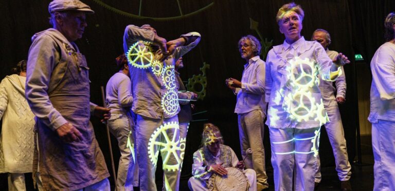 Een groep performers op een podium rondom een oplichtende mechanische installatie met grote verlichte tandwielen.