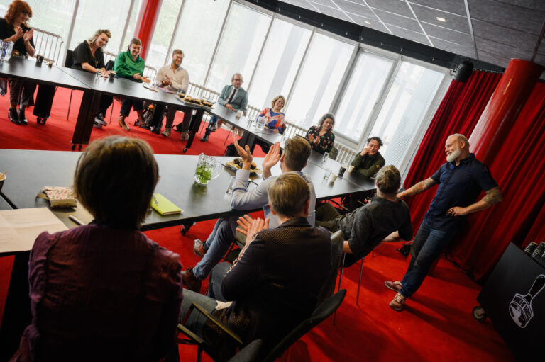 Een groep mensen zit rond een grote tafel in een vergaderruimte met veel natuurlijk licht.