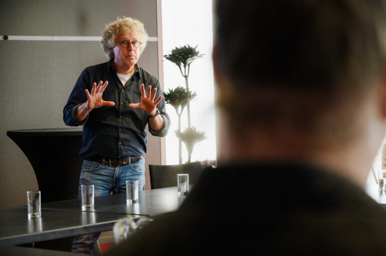 man met grijs krullend haar met donkergrijs overhemd staat met handen naast elkaar te vertellen voor de groep