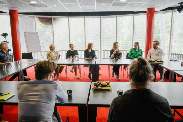 Een groep mensen zit rond een grote tafel in een vergaderruimte met veel lichtinval.
