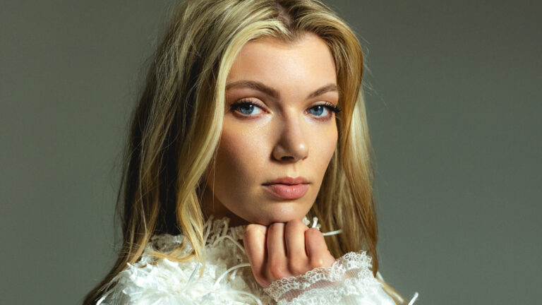 Blonde vrouw staat voor donkere achtergrond met witte kleding met veertjes