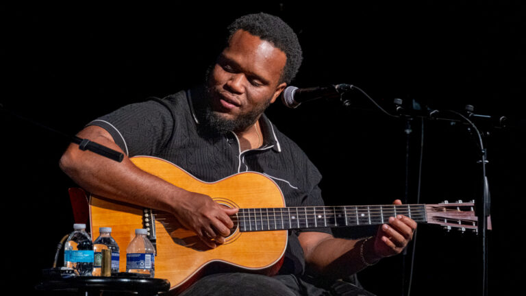 Man speelt akoestische gitaar op het podium met microfoons en waterflessen op de voorgrond.