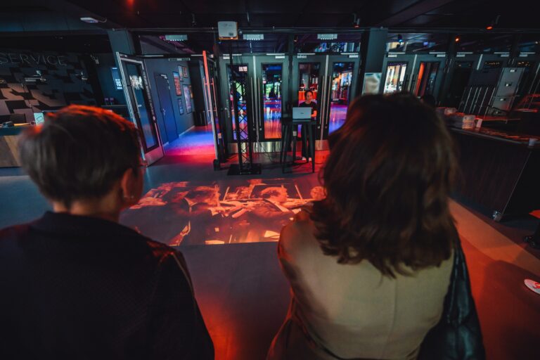 Twee mensen kijken naar een lichtprojectie op de vloer in een moderne, kleurrijke ruimte met displays.