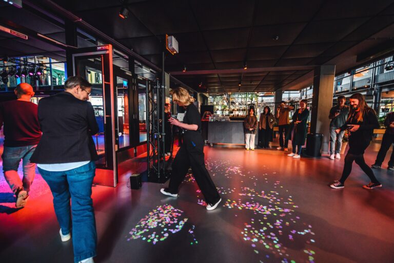 Een persoon in zwarte kleding loopt over de vloer, bedekt met kleurrijke confetti, terwijl anderen op de achtergrond toekijken.