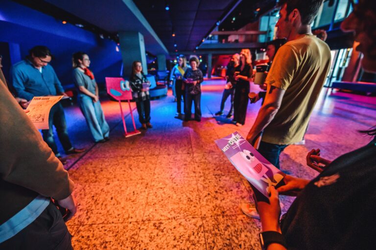 Een groep mensen verzameld in een kleurrijke ruimte, luisterend naar iemand die aan het woord is.