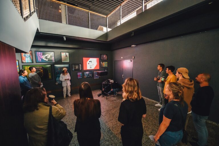 Een groep mensen luistert naar een presentatie in een kunstgalerie met een projector op de muur.