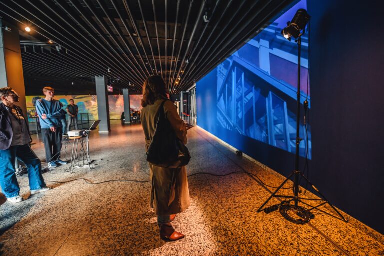 Een persoon bekijkt een groot scherm met een blauwe achtergrond in een moderne tentoonstellingsruimte.