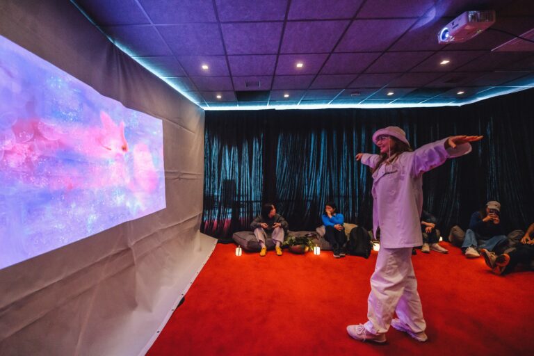 Een persoon in witte kleding staat voor een scherm met kleurrijke projecties, terwijl anderen op de achtergrond op de grond zitten.