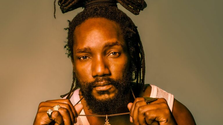 Man met donker haar en grote knot op zijn hoofd houdt de ketting die hij om heeft in de lucht.