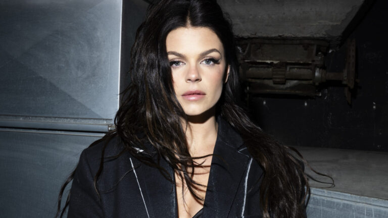 Davina Michelle kijkt gehuld in zwarte kleding serieus naar de camera met een donkere achtergrond.