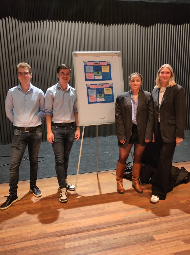 Vier mensen staan naast een bord met posterpresentaties in een zaal.
