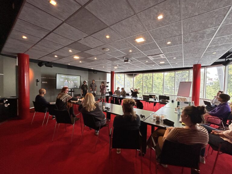 groep mensen kijkt naar de presentaties van de deelnemers en zitten aan een grote tafel