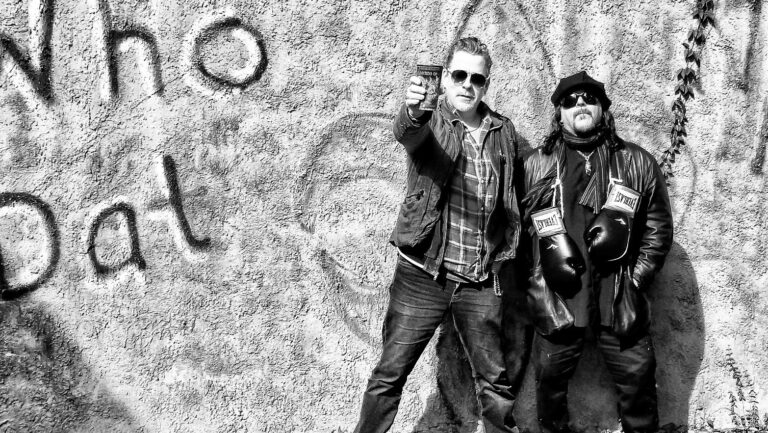 Twee mannen poseren voor een muur met graffiti waarop 'Who Dat' staat, waarbij de ene man een drankje omhoog houdt en de andere man een leren jas en bokshandschoenen draagt.