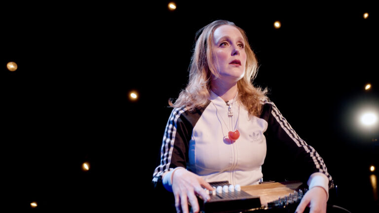 Een blonde vrouw staat achter een keyboard op een donker podium met kleine lichtjes op de achtergrond.