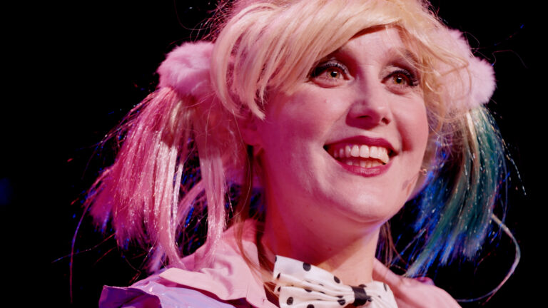 Een vrouw met blonde haren glimlacht op een podium onder roze verlichting.