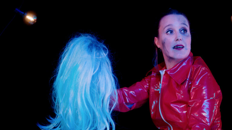 Twee personen op een donker podium, één met felblauw haar en één in een rode outfit met een verraste uitdrukking.