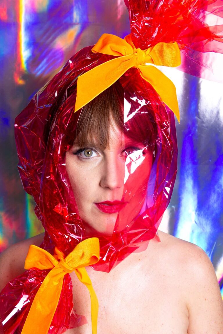 Een vrouw met een glanzende, rode plastic hoed en een gele strik poseert tegen een kleurrijke, reflecterende achtergrond.