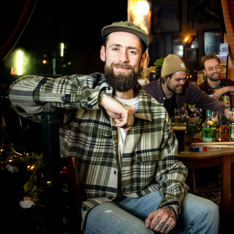 Man met baard in café, draag geruit overhemd, poseert ontspannen, gezellige sfeer, vrienden op achtergrond.
