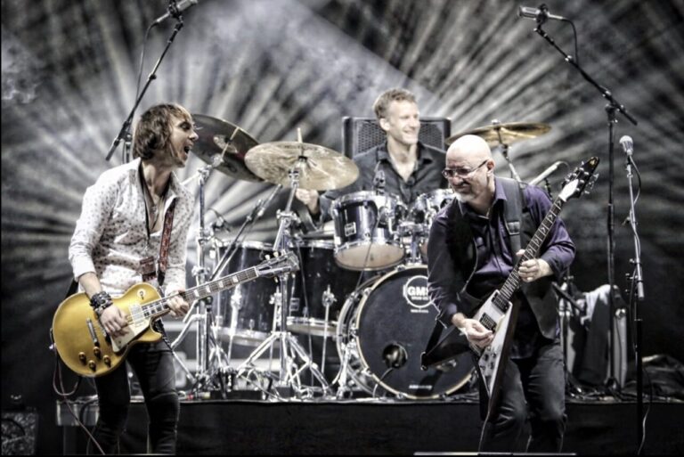 Drie leden van Wishbone Ash spelen enthousiast live op het podium, met dramatische verlichting op de achtergrond.