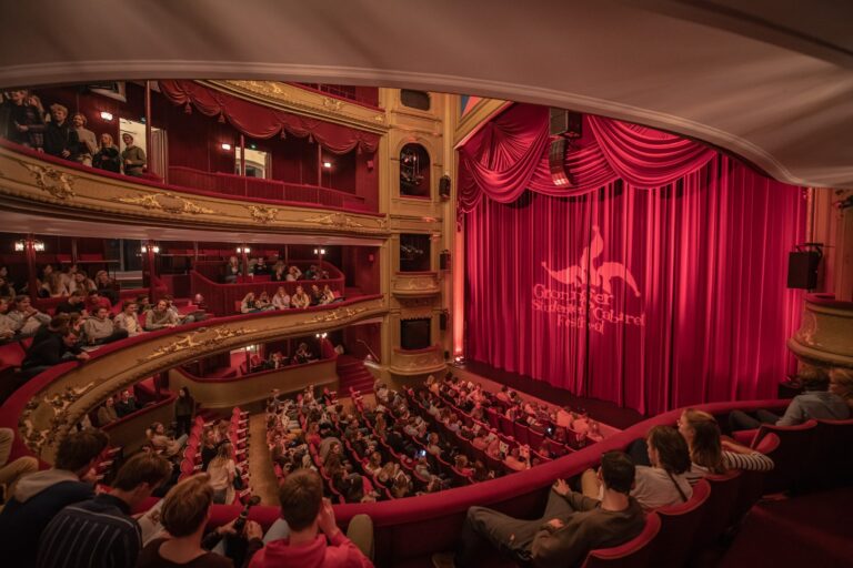 Een vol theater met roodfluwelen zitplaatsen en gordijnen, waarbij het publiek wacht op de voorstelling, met de naam