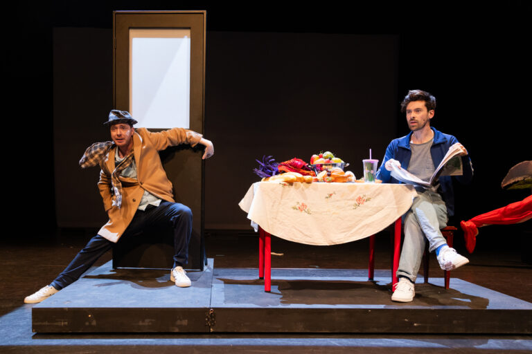 Twee mannen op een theaterpodium, waarvan één zittend aan een tafel en de ander leunend tegen een deur.