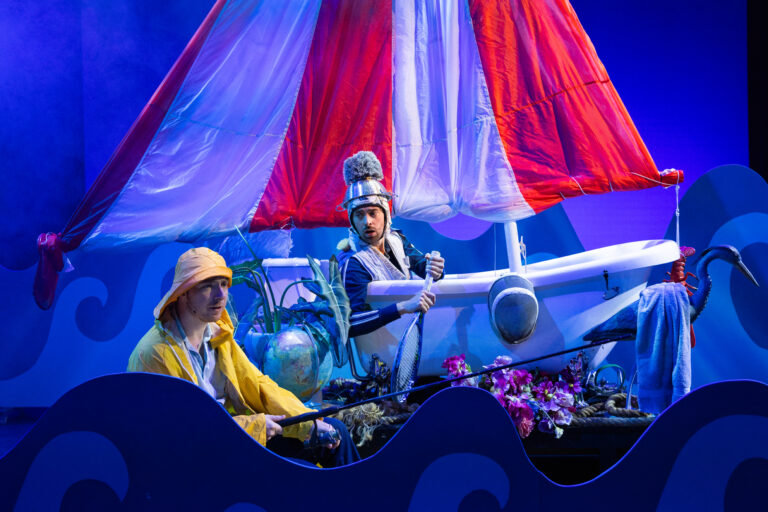 Twee acteurs zitten in een speelgoedboot met een rood-witte zeildoek, omringd door blauwe golven en een globe.