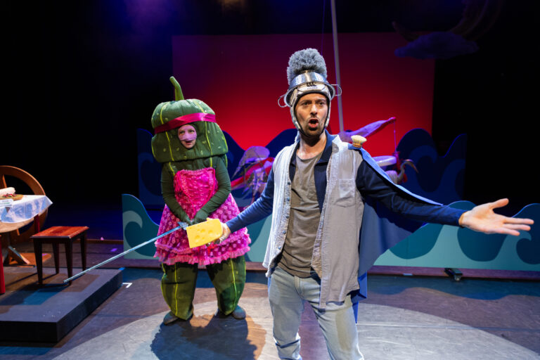 Twee acteurs op het podium, waarvan één in een kostuum van een groente en de ander gekleed als een personage met een helm en zwaard, staan in een theatrale setting.