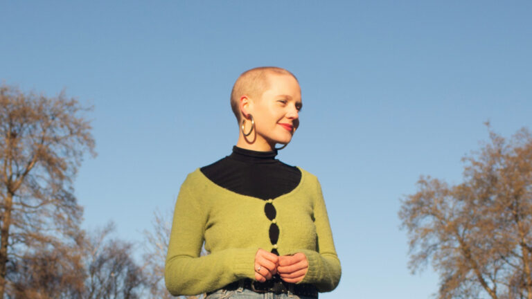 Singer-songwriter in a green cardigan standing outdoors, smiling with a clear sky background.