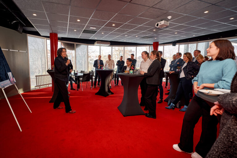 Groep mensen luistert naar een spreker in een zaal met rode vloerbedekking.