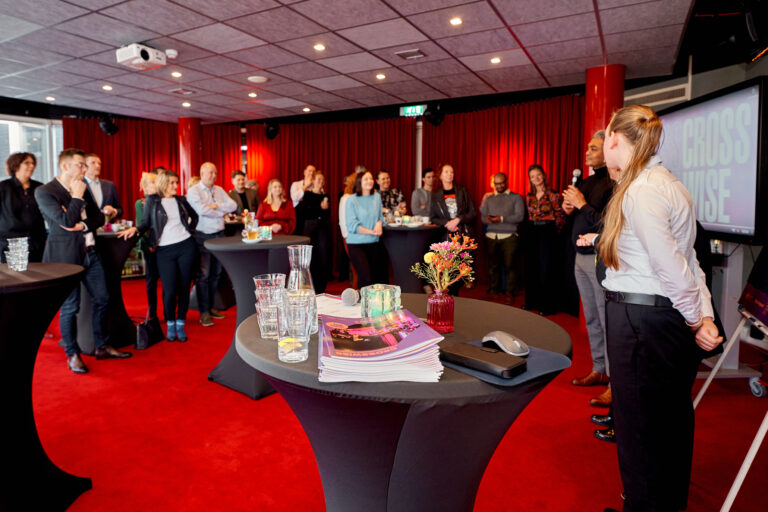 Een groep mensen staat in een kamer met rode vloerbedekking en rode gordijnen, met staande tafels in het midden.